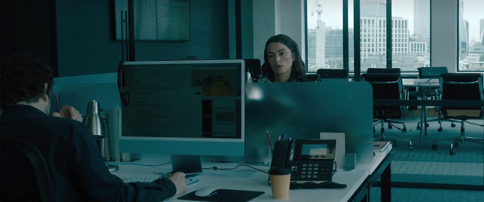 Backplates for film & TV by Will Pearson. The image is a still from The Woman in Cabin 10 © Netflix. It shows a woman (actress Keira Knightley) sitting at a desk in an office setting with the London skyline visible through the meeting room windows.