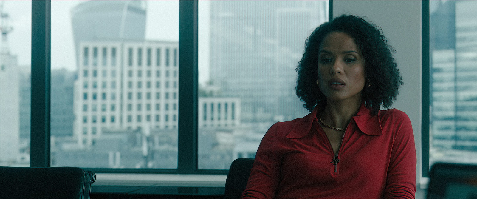Backplates for film & TV by Will Pearson. The image is a still from The Woman in Cabin 10 © Netflix. It shows a woman in a red top sitting in an office setting with the London skyline visible through the office windows.